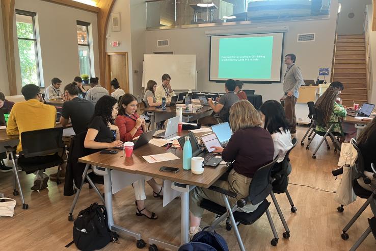 A group of training participants sit at desks in front of an instructor who stands at the front of the room by a projector and training slides.