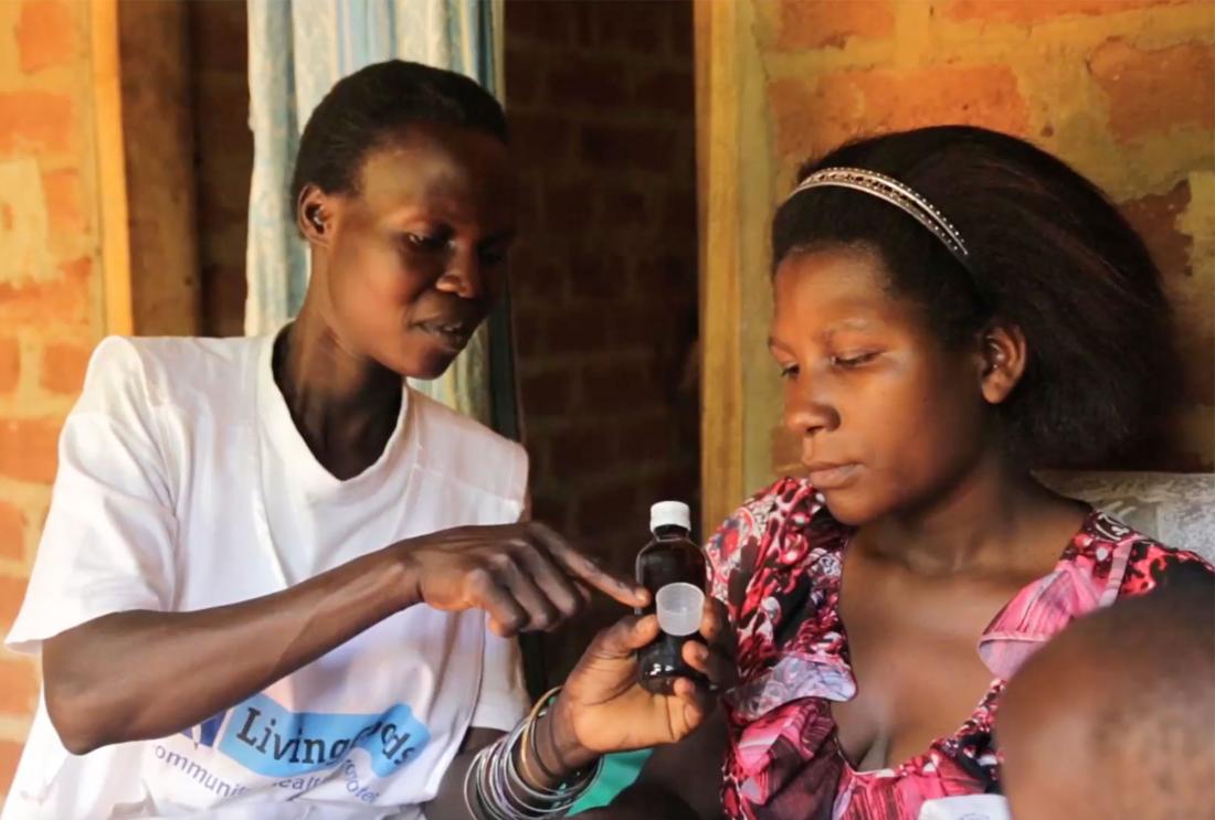 Woman in Living Goods uniform points at medicine bottle while mother of newborn watches