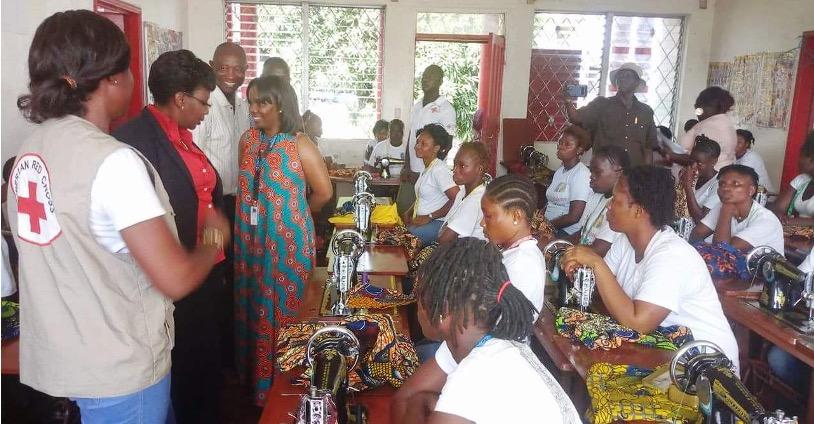Women in Liberia sit at tables with sewing machines and cloth in front of them. Instructors, including one wearing a vest from the Liberian Red Cross, smile at the front of the room.