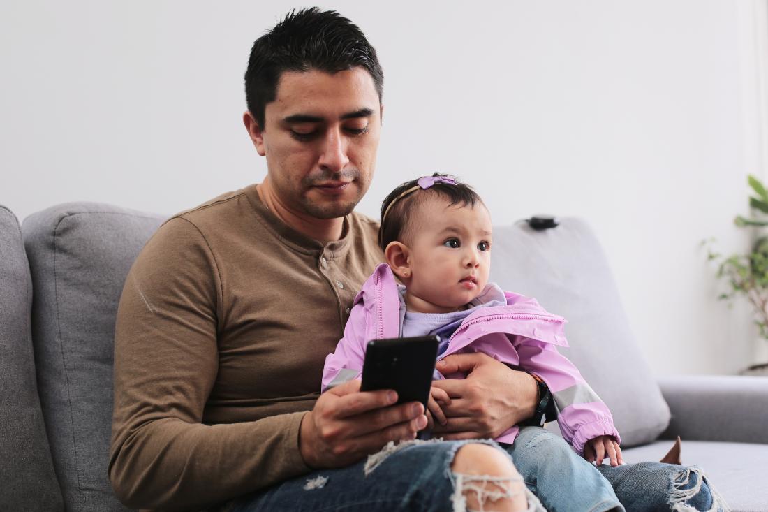 Young dad holding baby in his arms while using cell phone