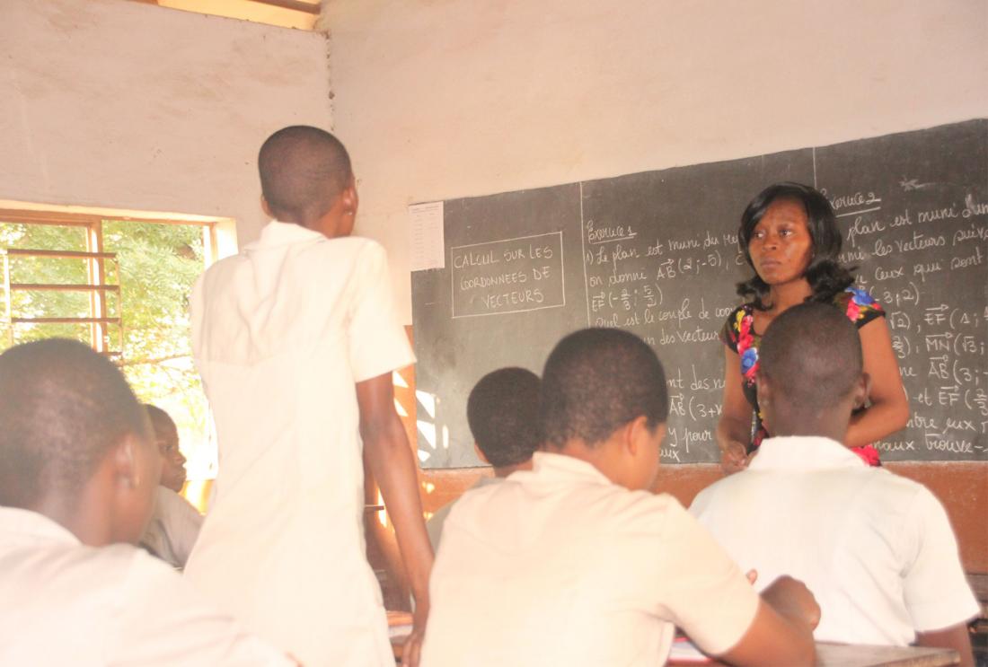 Woman stands in front of classroom chalkboard while student stands facing her