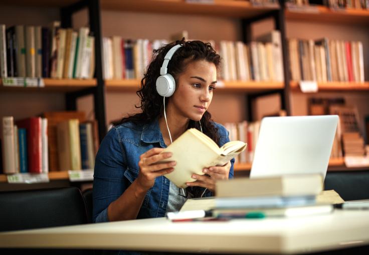 A woman wearing headphones and holding a book studies in a library and looks at a laptop.