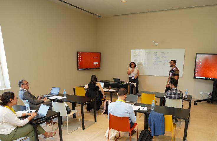 Training participants sit in a classroom, two teachers stand in the front and are smiling.