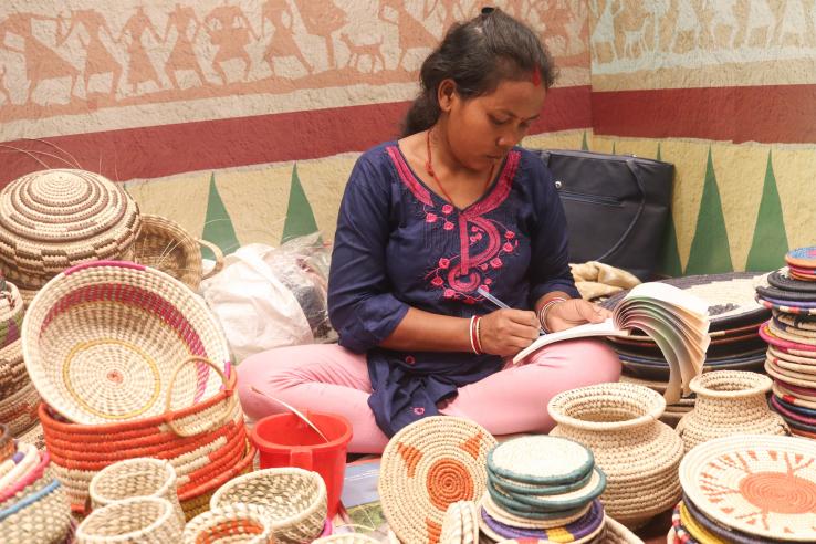 A woman writes on a notepad, seated amongst basket merchandise for Microfinance J-PAL RCT