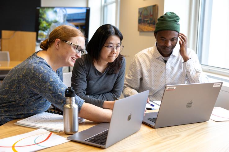 Three DEDP MicroMasters students sit around a table and discuss coursework with their laptops open.