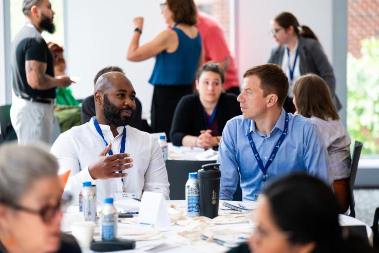 people chatting with their neighbors at tables during convening, with focus on two men in midground