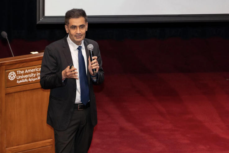 A man in a suit stands on a red stage and speaks into a handheld microphone