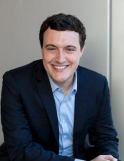 Headshot of Joshua Dean wearing a blue shirt and black coat