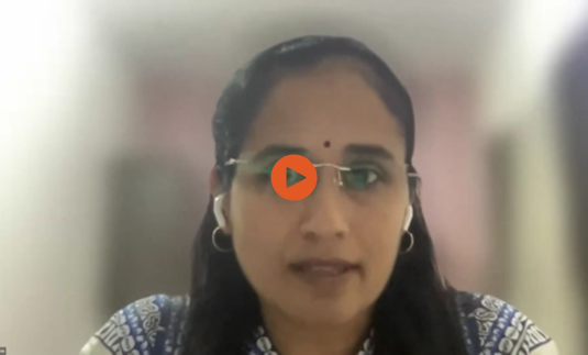 A woman with glasses and earbuds is speaking during a virtual meeting. She has long dark hair, a bindi on her forehead, and is wearing a patterned blue top. A blurred indoor background is visible behind her. A play button overlay is centered on the image, indicating this is a video still.