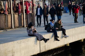 Three women sitting and relaxing on stone ledge by water, looking at phones