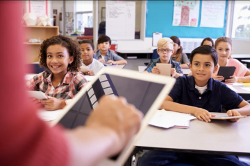 children sit at a desk in front of a teacher with an ipad 