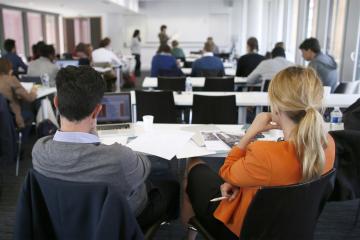 Two people sitting at a desk, observing a lecturer.