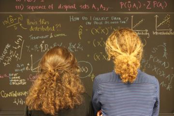 Two individuals looking at a chalk board with formulas and notes written on it.