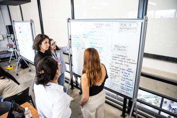 Three people stand around a white board and point to it.