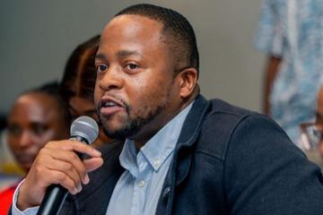 Takudzwa Mutize speaks while sitting down, wearing a suit, and holding a microphone